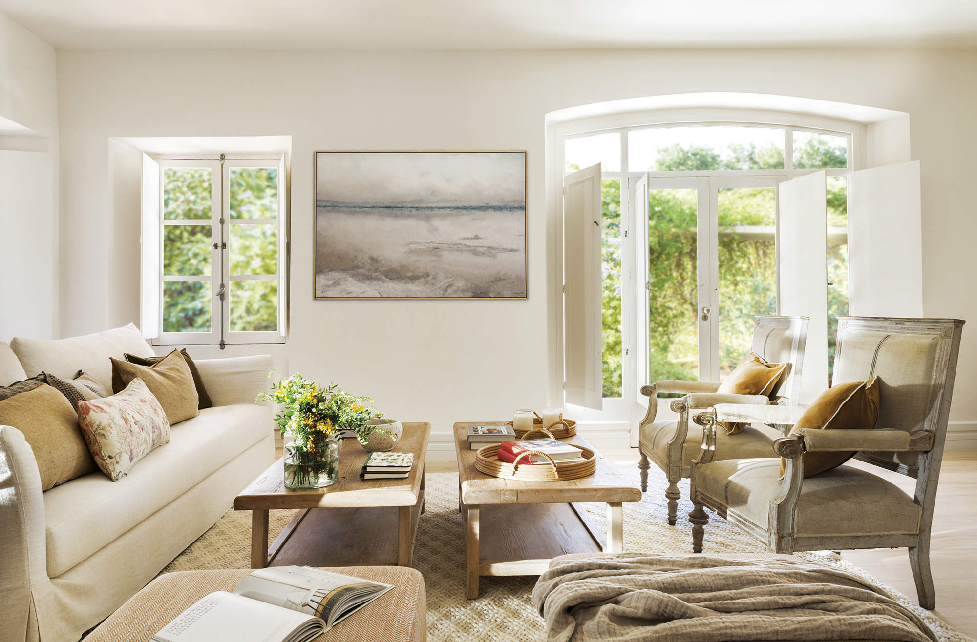 salón con sofá blanco y cojines mostaza, dos mesas de centro de madera, cuadro, ventanal al jardín, butacas de madera tapizadas